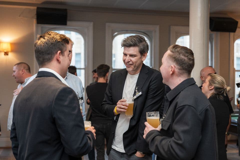 Three guests in suits talking over beers by the windows of a function room
