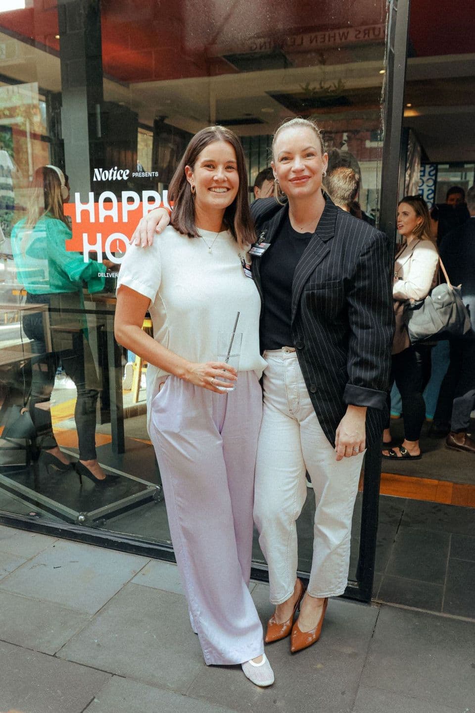 Two guests on the footpath outside a Notice Happy Hour, the poster in the window behind them