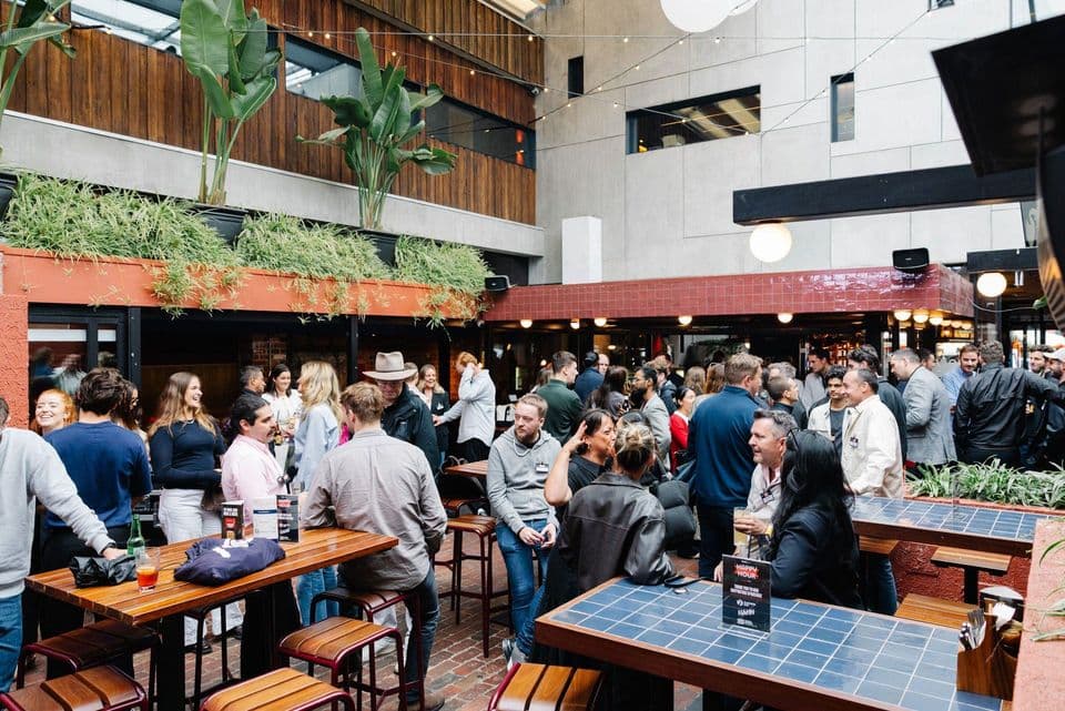 A full courtyard of guests standing at long timber tables at a Notice Happy Hour