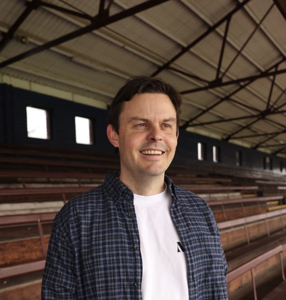 Matthew Hughes in a checked shirt, smiling, in an empty timber grandstand
