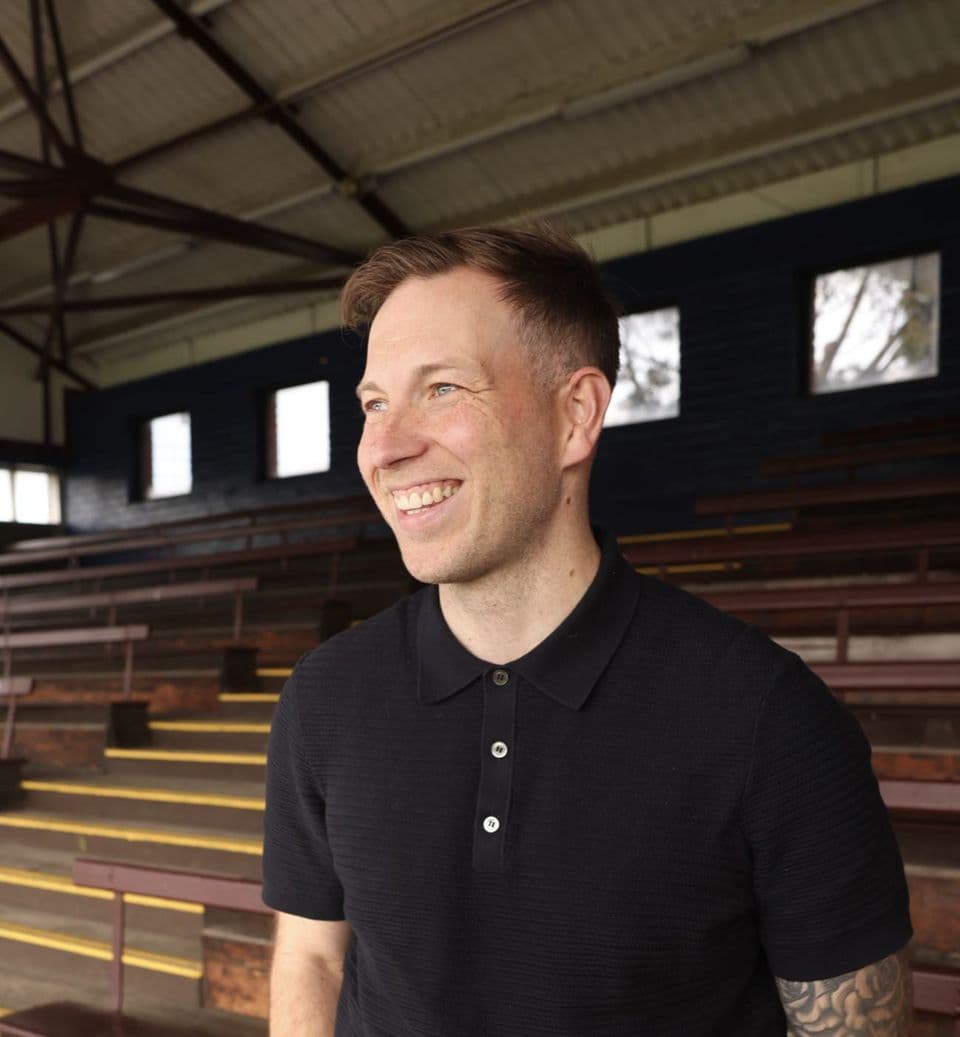 Tom Meredith in a black polo shirt, laughing, in an empty timber grandstand